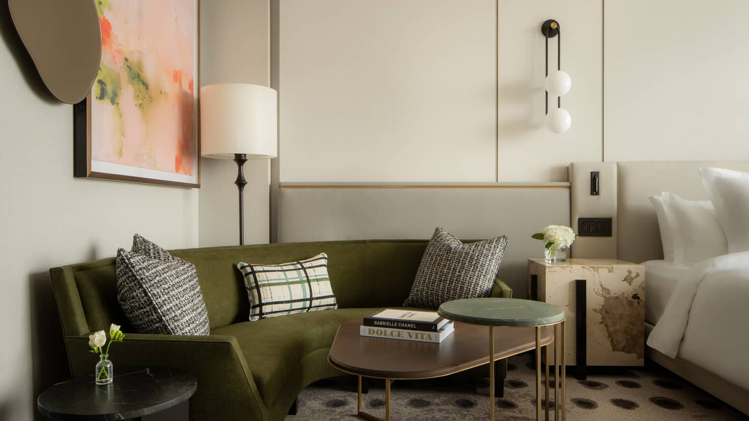 Olive green velvet sofa in a suite seating area with a black side table, patterned pillows, and a curved wooden coffee table stacked with designer books