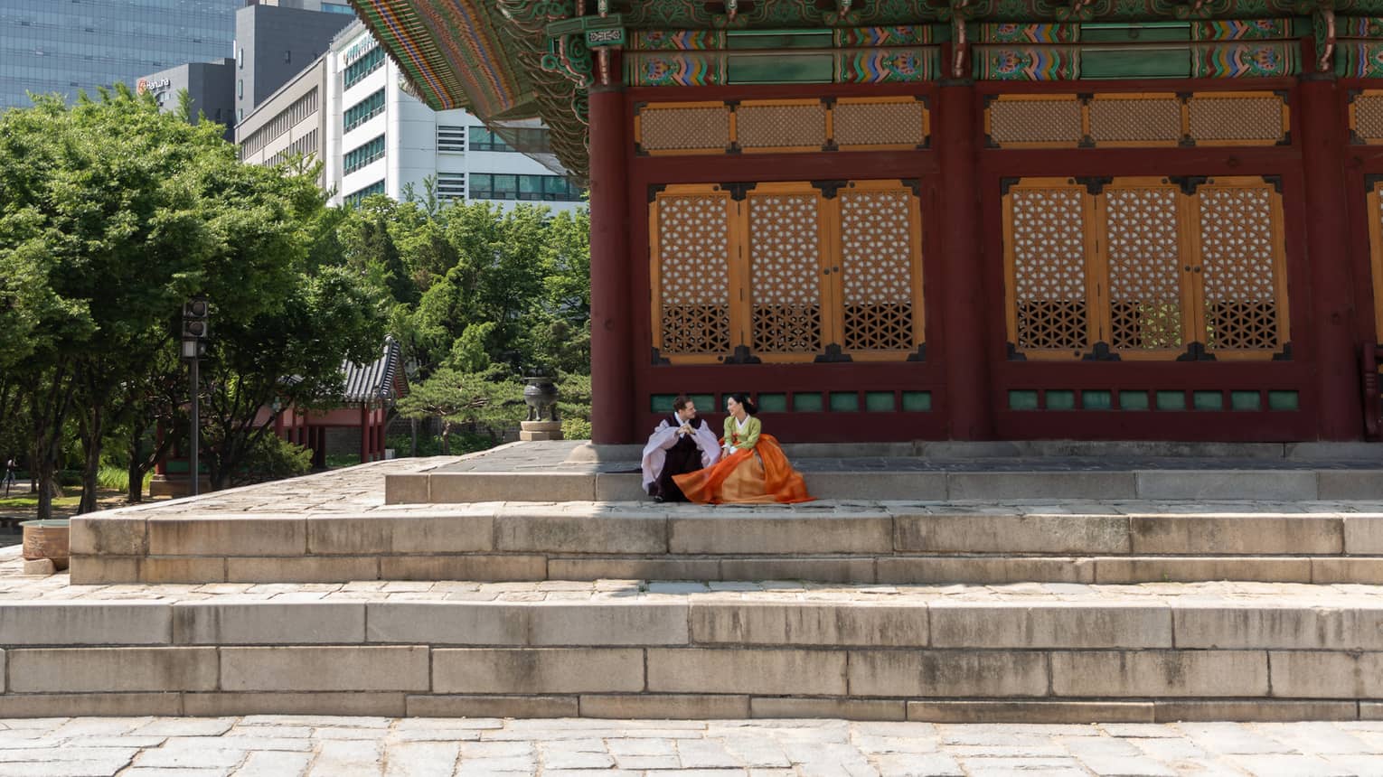 A couple in traditional Korean dress are seated on stone steps under the colourful roof awning of a tree-lined palace.