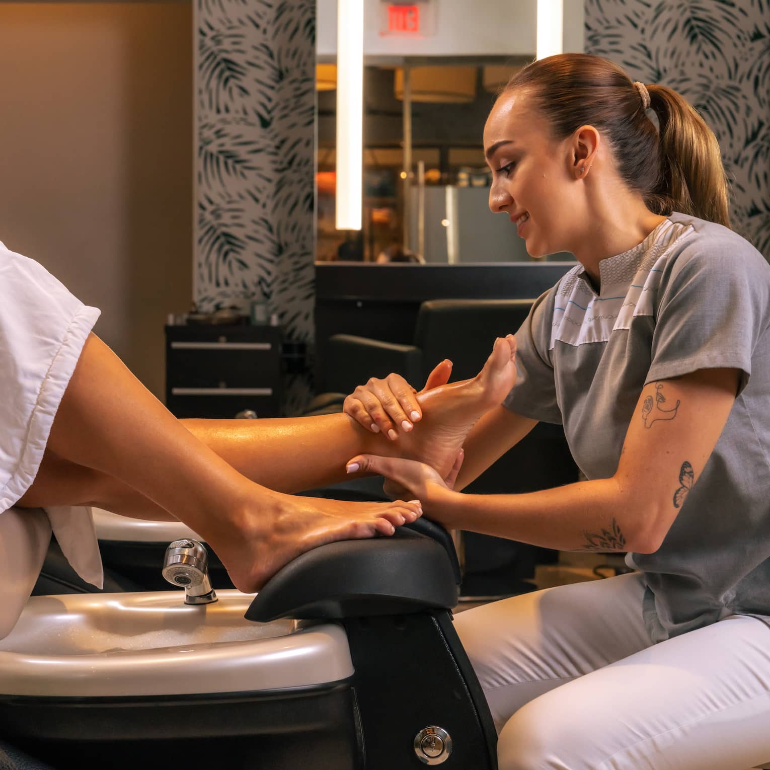 Pedicurist wearing grey shirt and white pants rubs the foot of a client sitting in a pedicure share and wearing a white robe