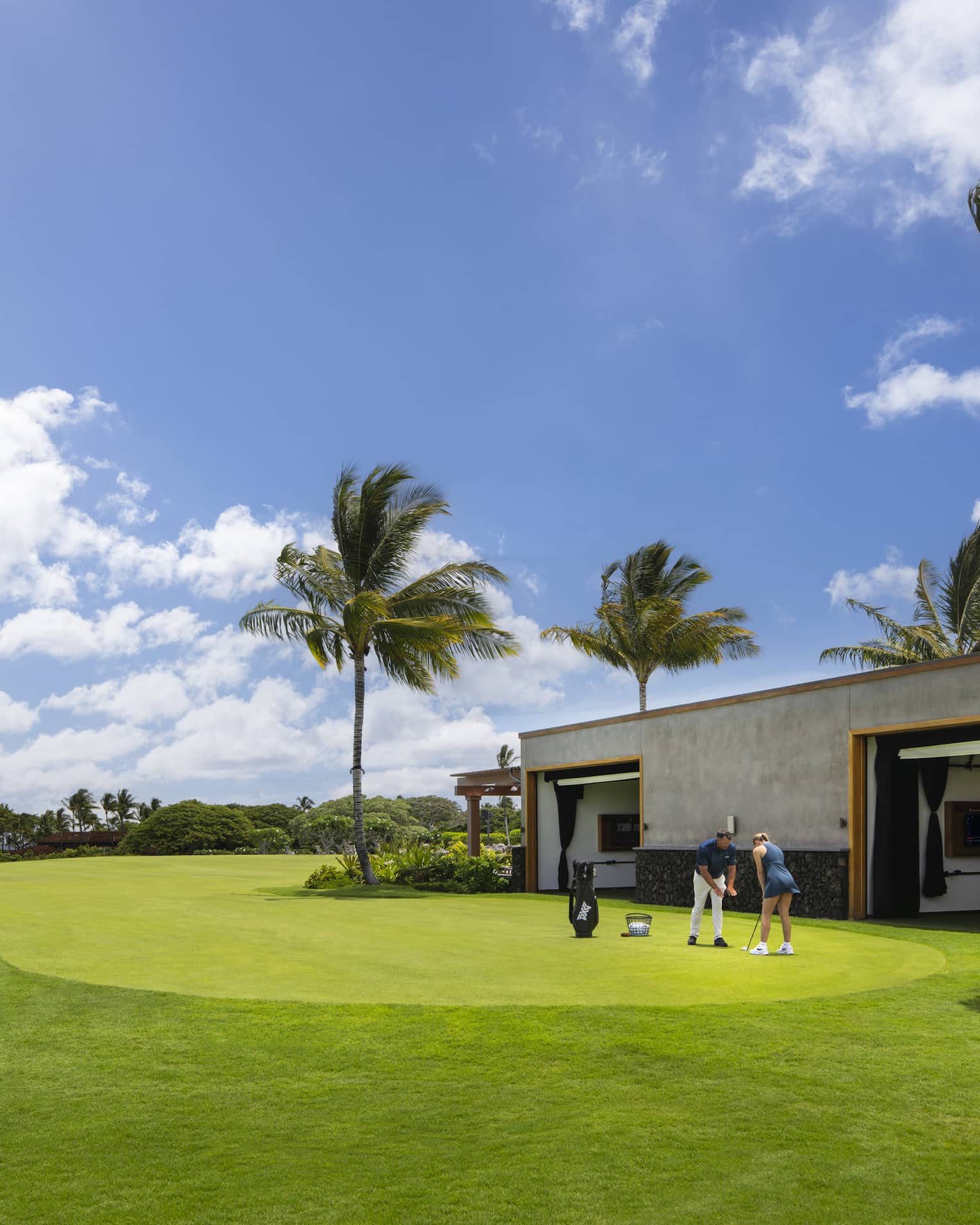 A woman receives an individual golf lesson on a golf course outside a building with palm trees.