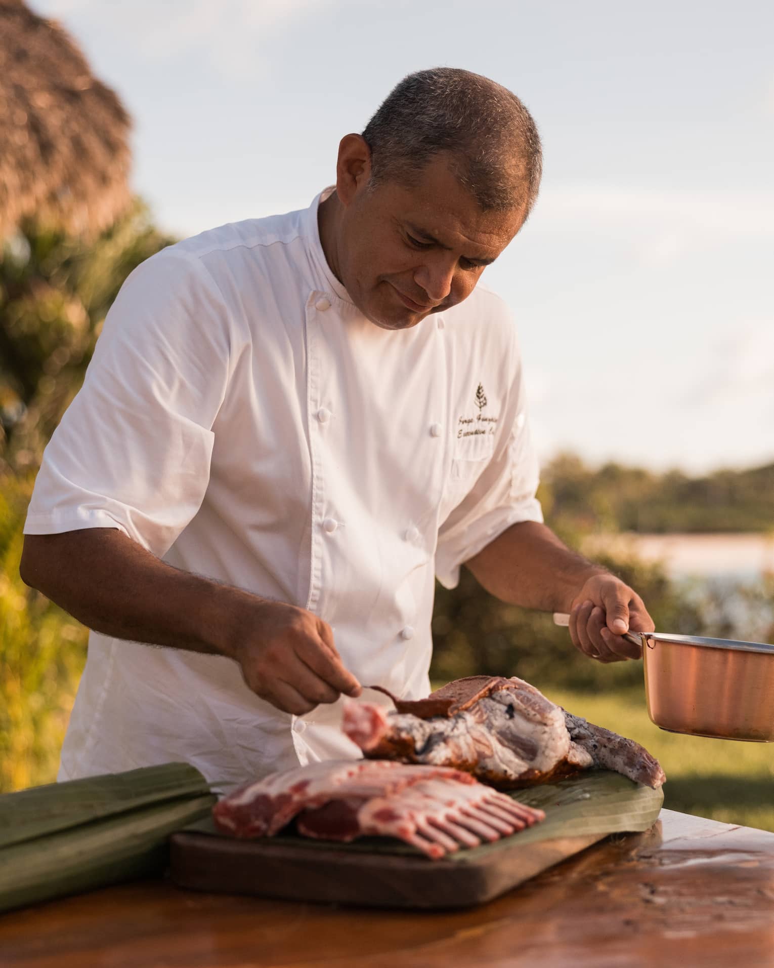 Chef in white chef’s coat, standing at an outdoor table, holding a copper pot in one hand and seasoning meat with the other