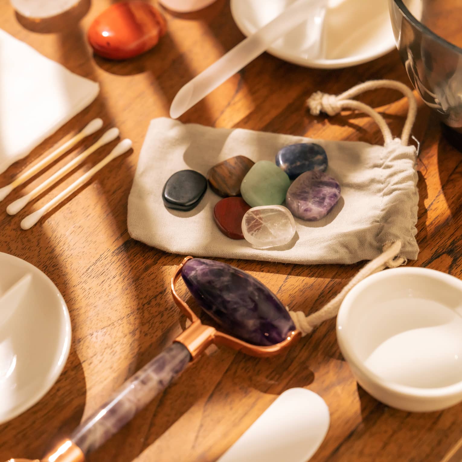 Face roller, gemstones, cotton swabs and a variety of small white bowls and other instruments sit on a sun-lit wooden table