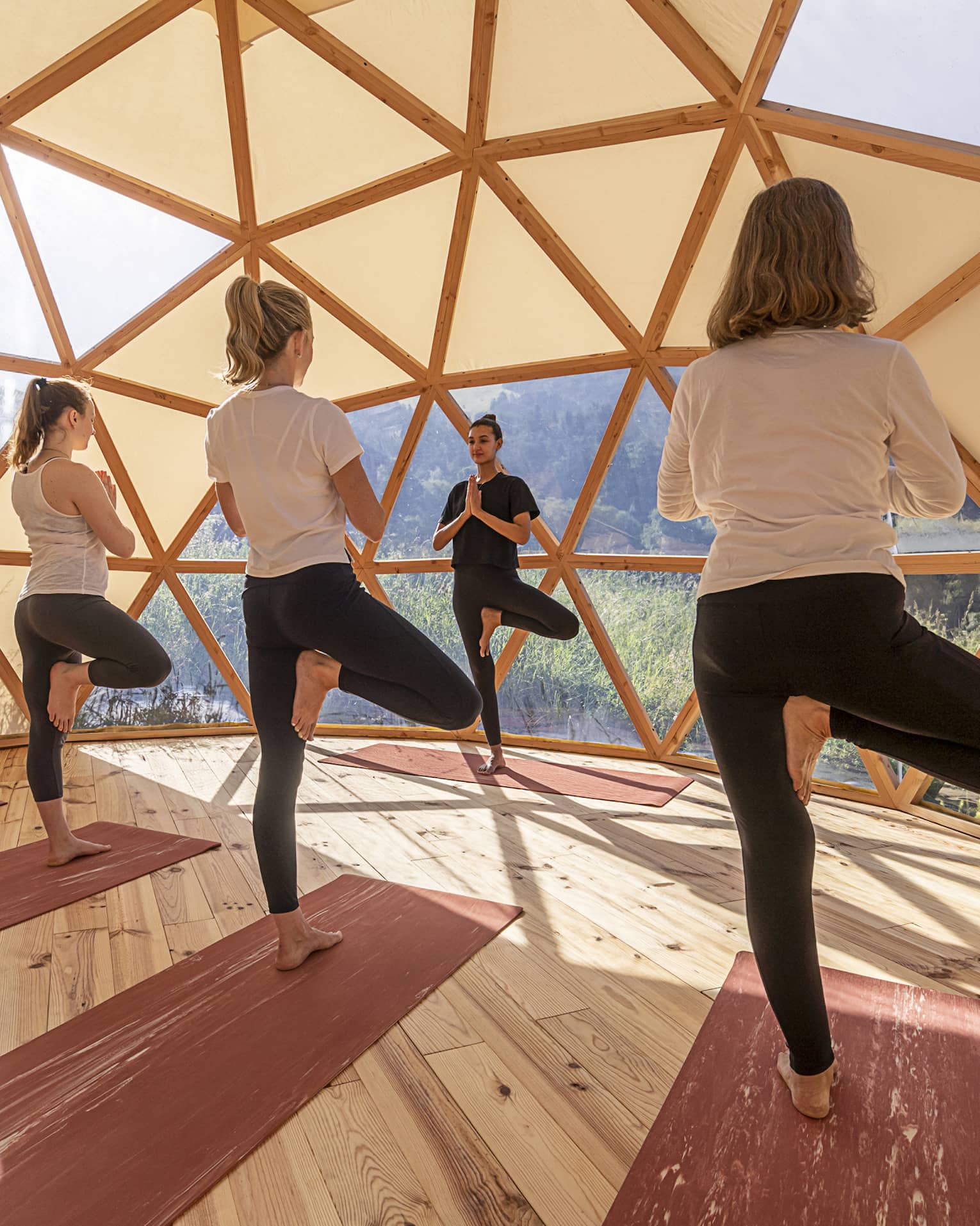Three women standing in crane pose facing yoga instructor inside eco-friendly geodesic dome