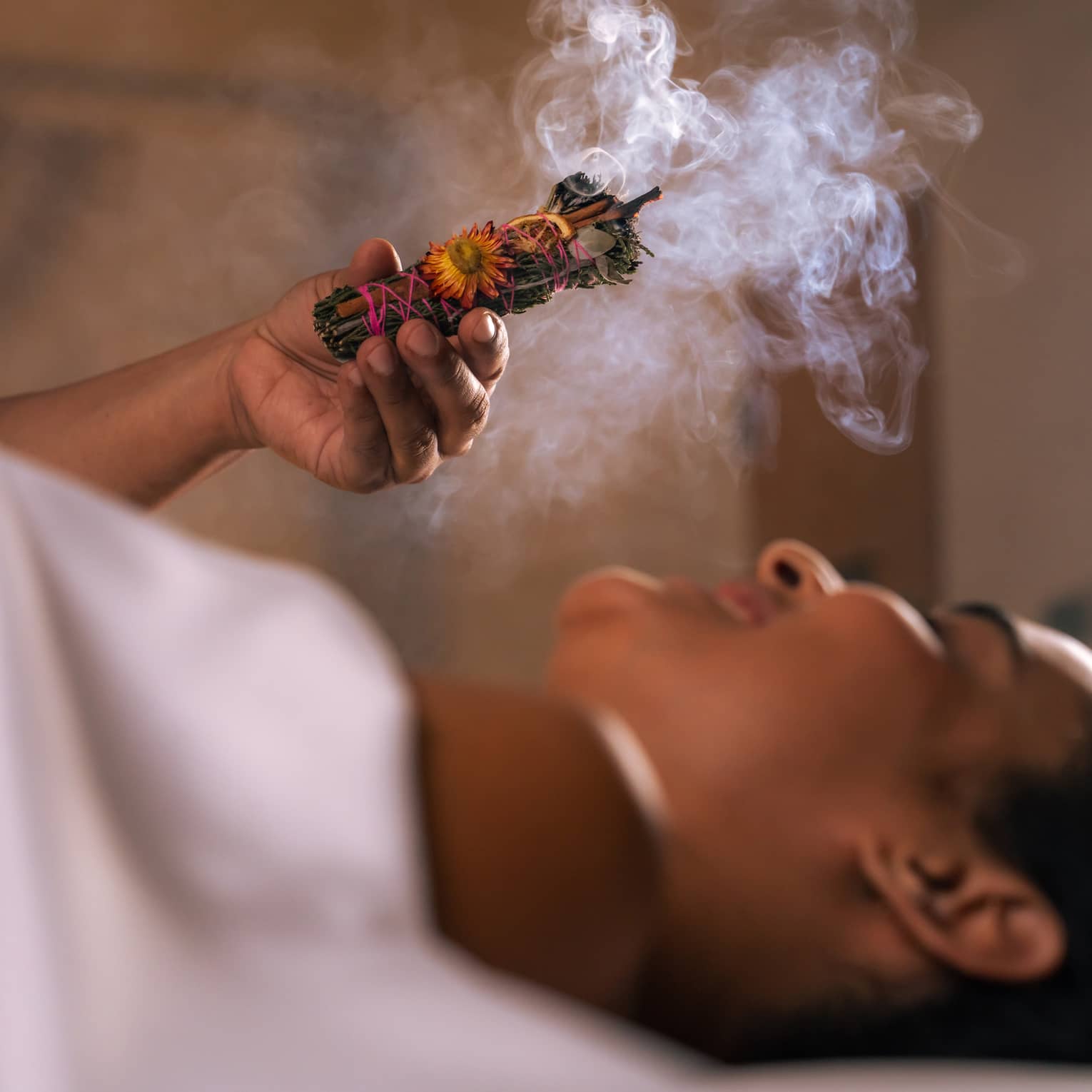 Person holds a smudge stick with smoke billowing over another person's head as they lie under a white sheet