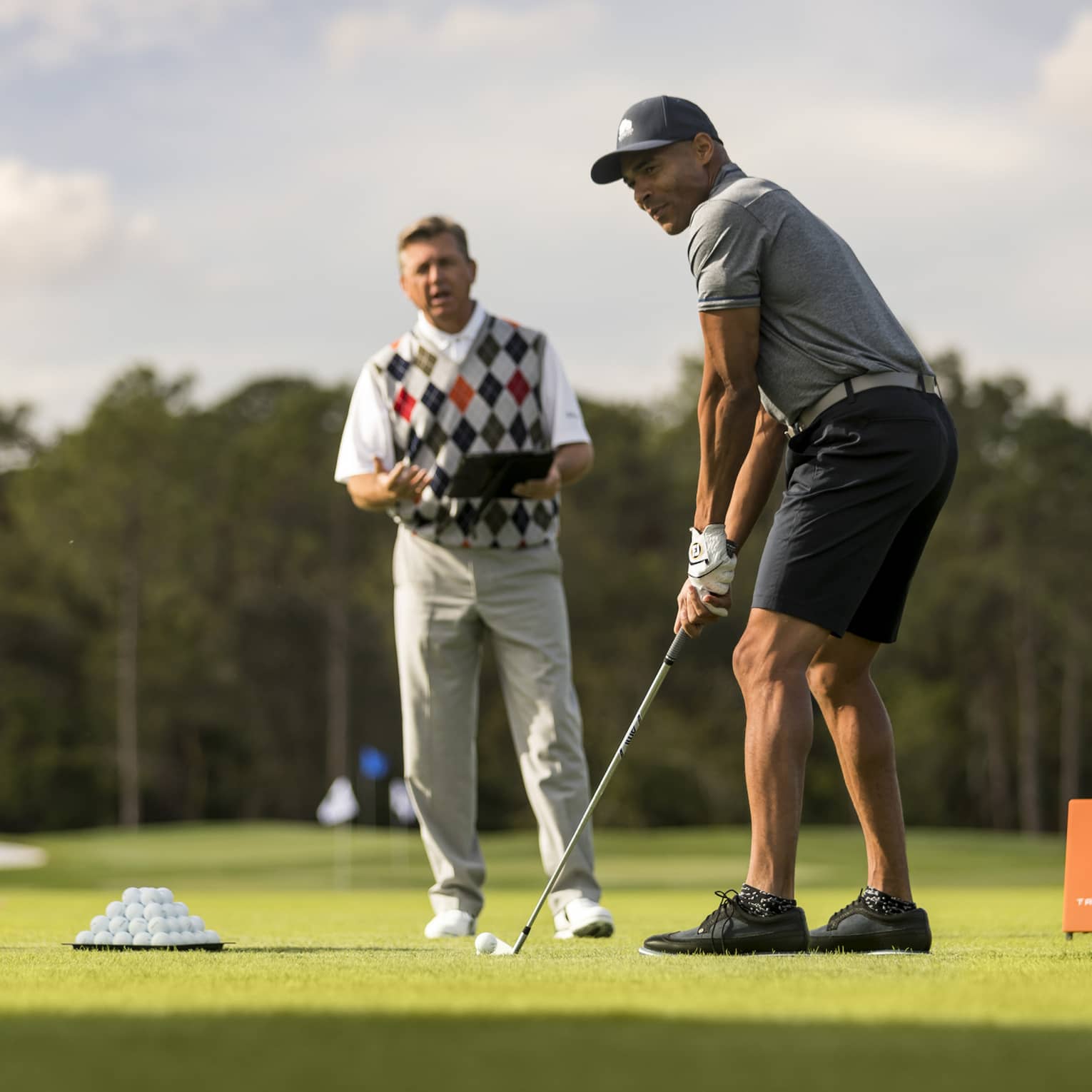 Against a clear sky, a golfer tees up, a tray of balls and a stand of clubs nearby. An instructor stands in the background.