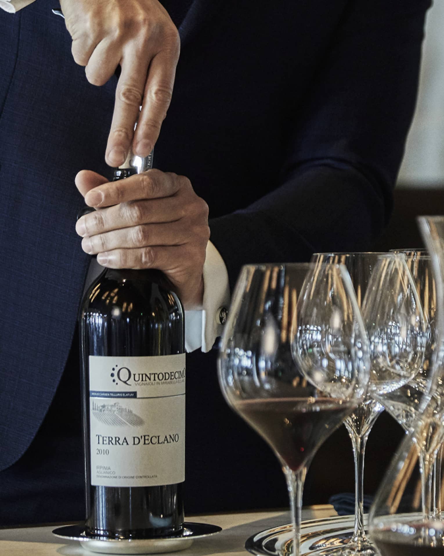 A person opening a bottle of wine, with wine glasses and additional wine bottles beside it