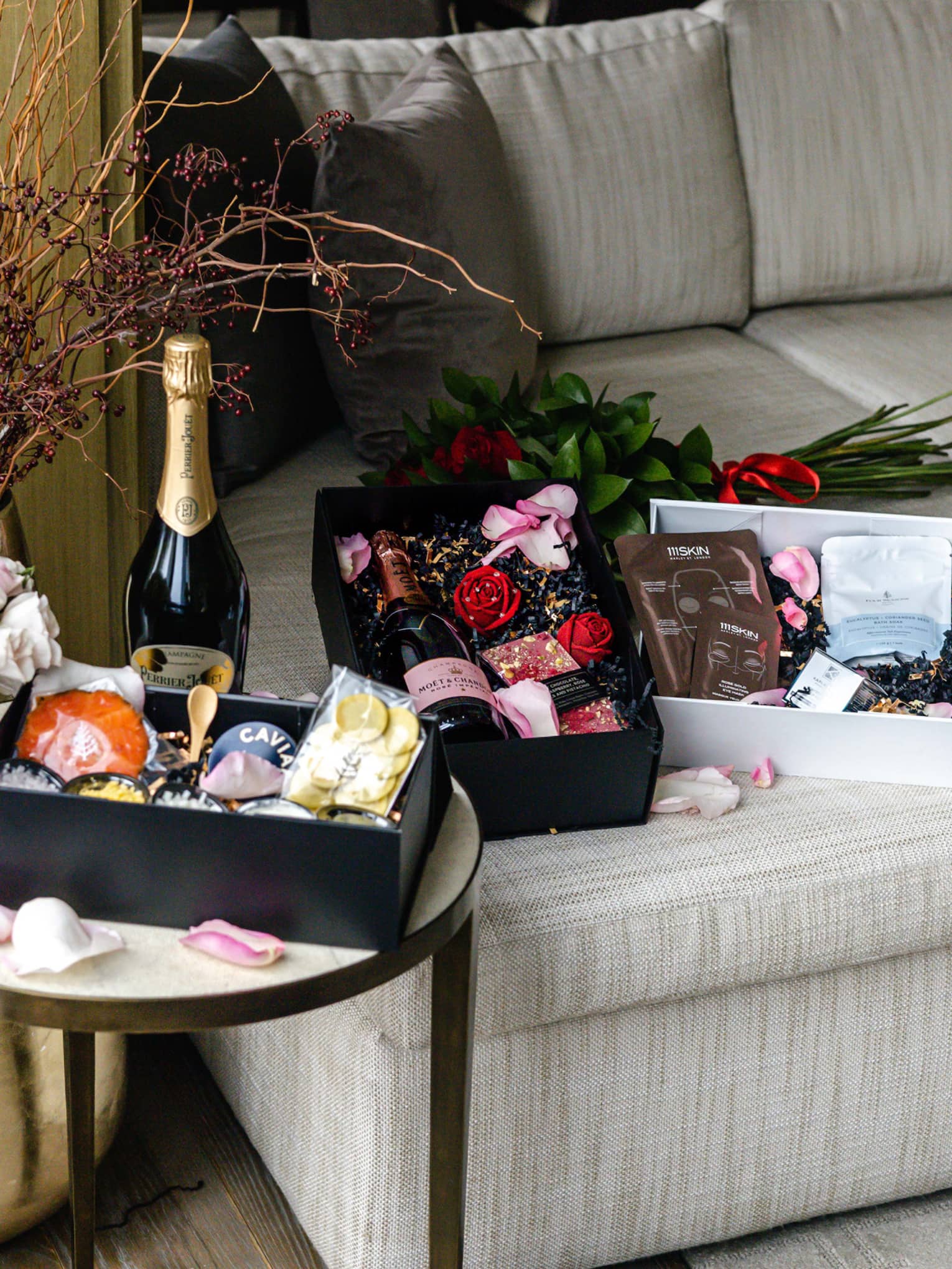 Three Valentine's Day gift boxes, a bottle of champage and two bouquets of flowers are shown on a small round table and a grey L-shaped couch
