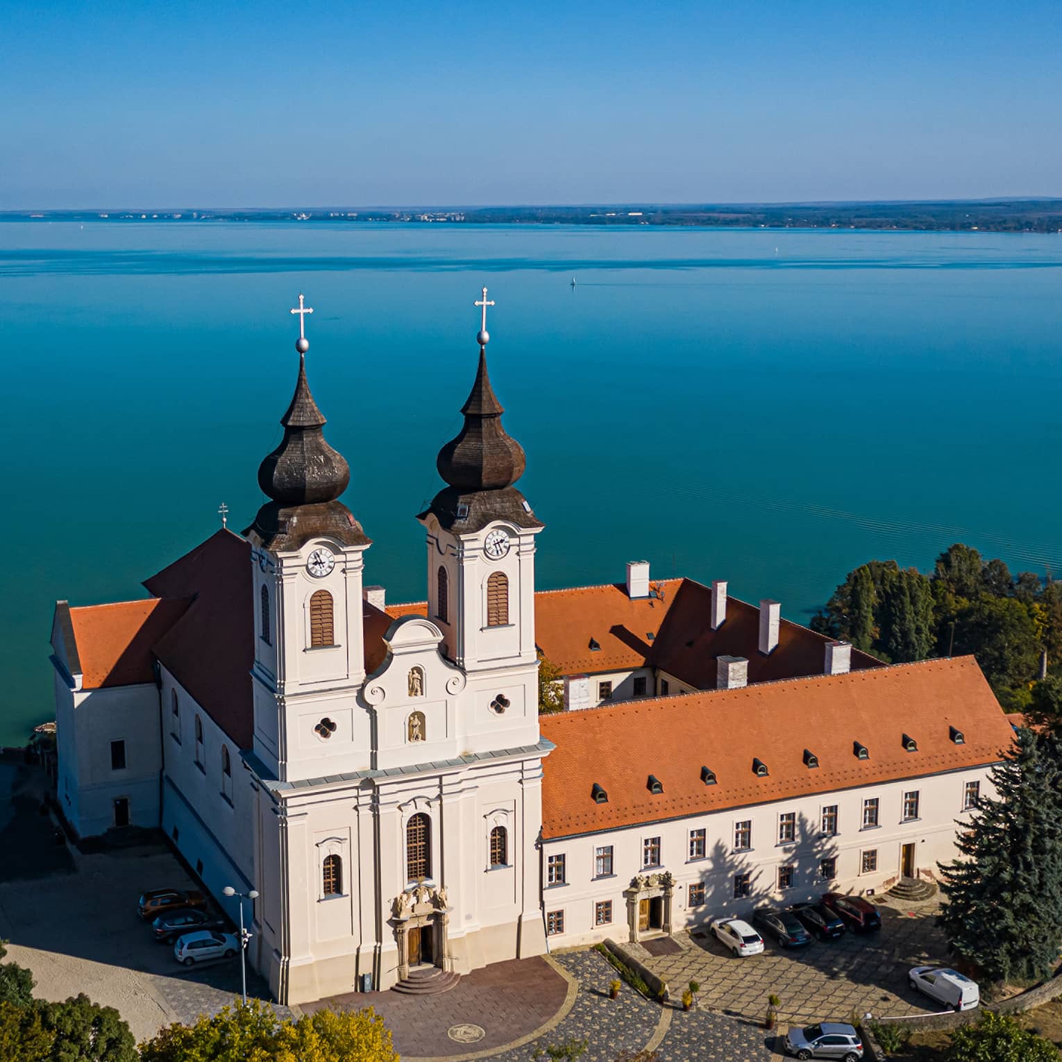 A Benedictine monastery, with white walls and a red tiled roof, sitting beside a large blue lake and surrounded by trees.