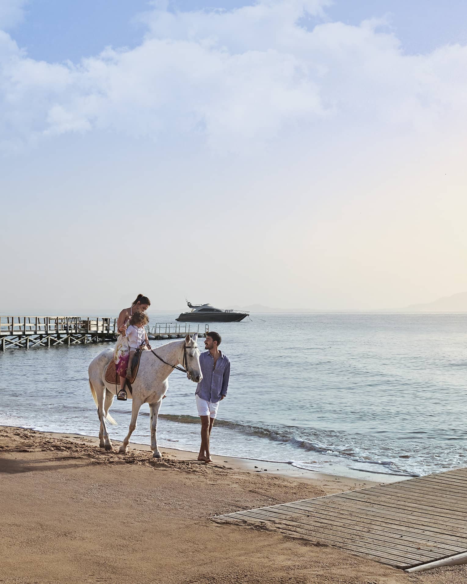 An adult and child on a white horse on a beach, led by another guest, with a wooden pier and a yacht visible in the distance.