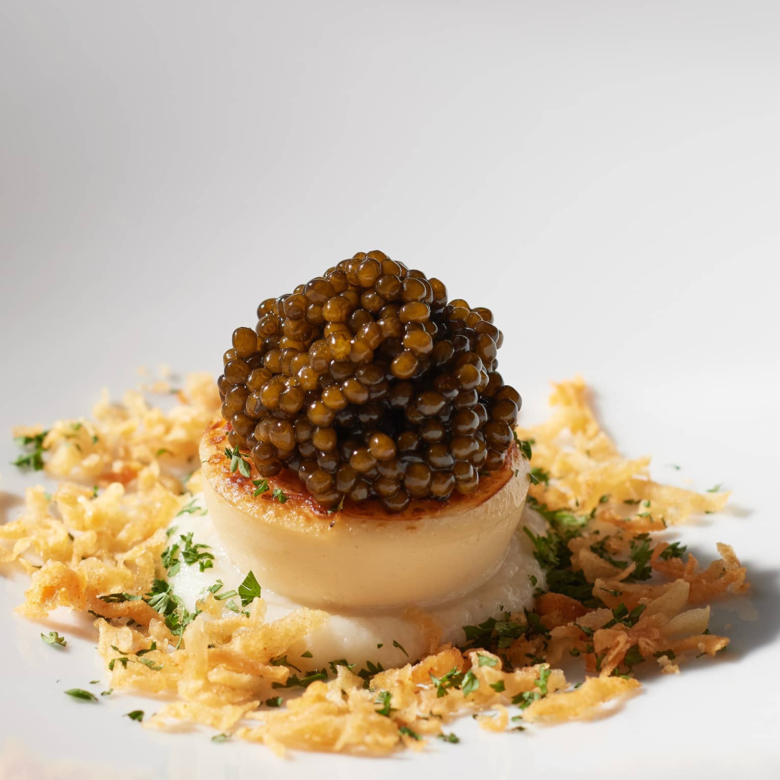 Round white plate with single scallop topped with caviar, surrounded by breadcrumbs and a smattering of green herbs