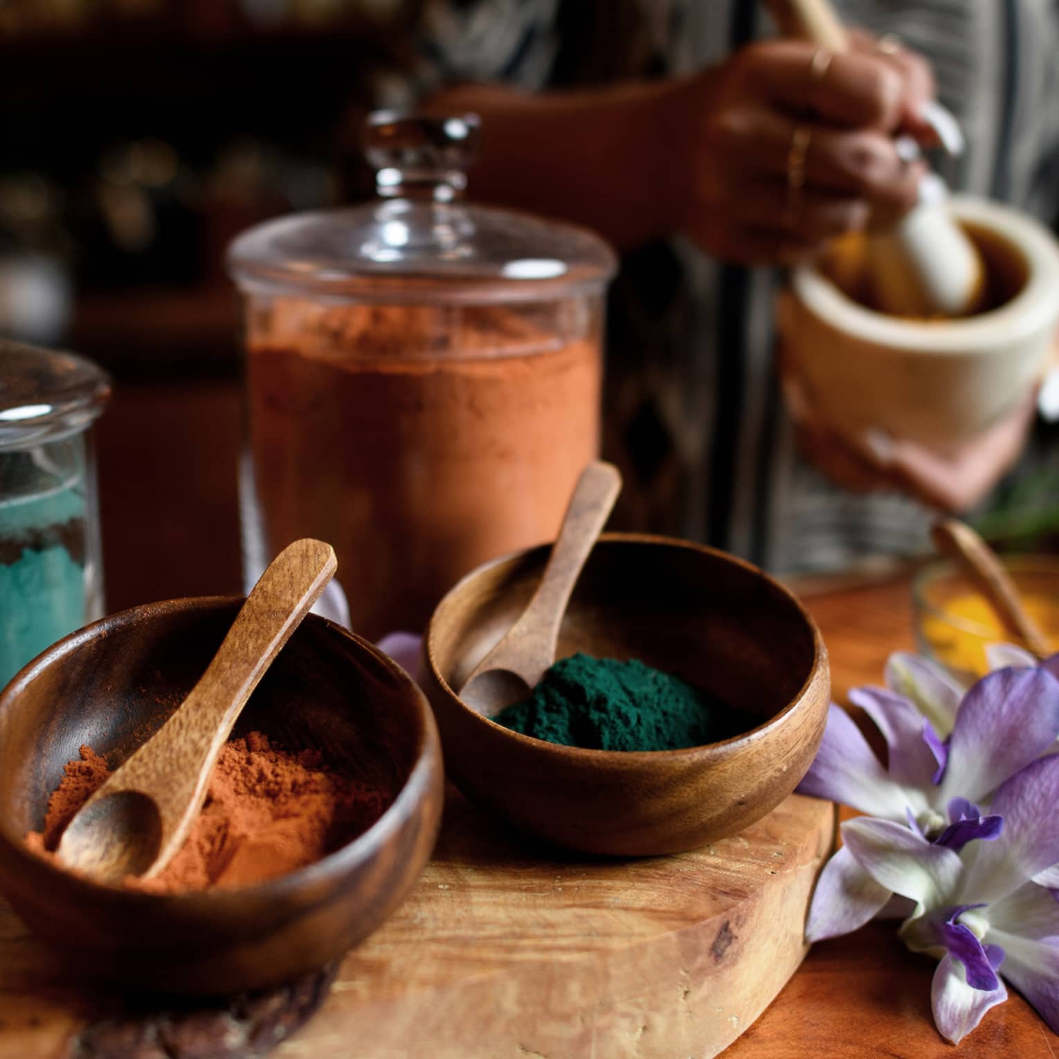 Small wood bowls and spoons beside glass jars filled with colourful powders, tropical flower