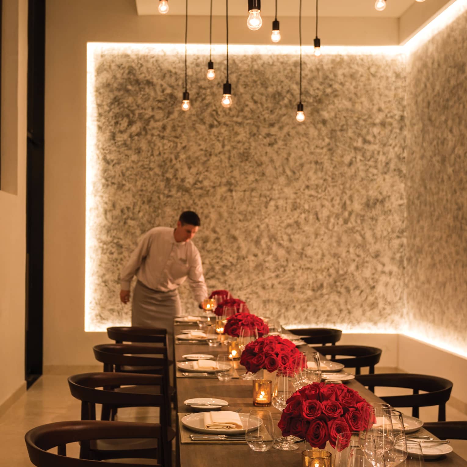 A server lights a candle at the far end of a long table artfully arranged with vases of red roses and set for a large group.