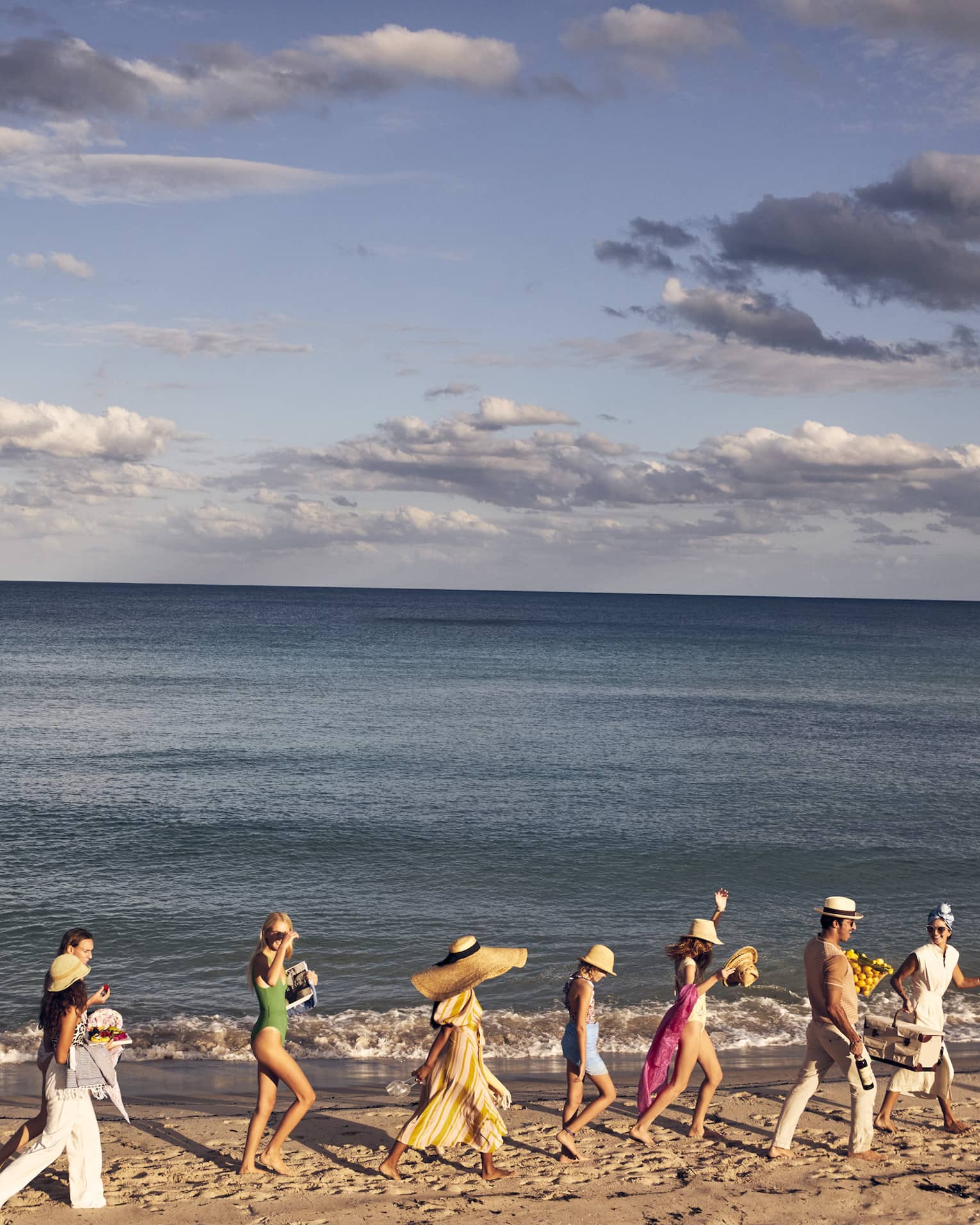 A group of people walking along a sandy beach by the ocean, wearing summer clothing and hats, with a backdrop of a calm sea and partly cloudy sky.
