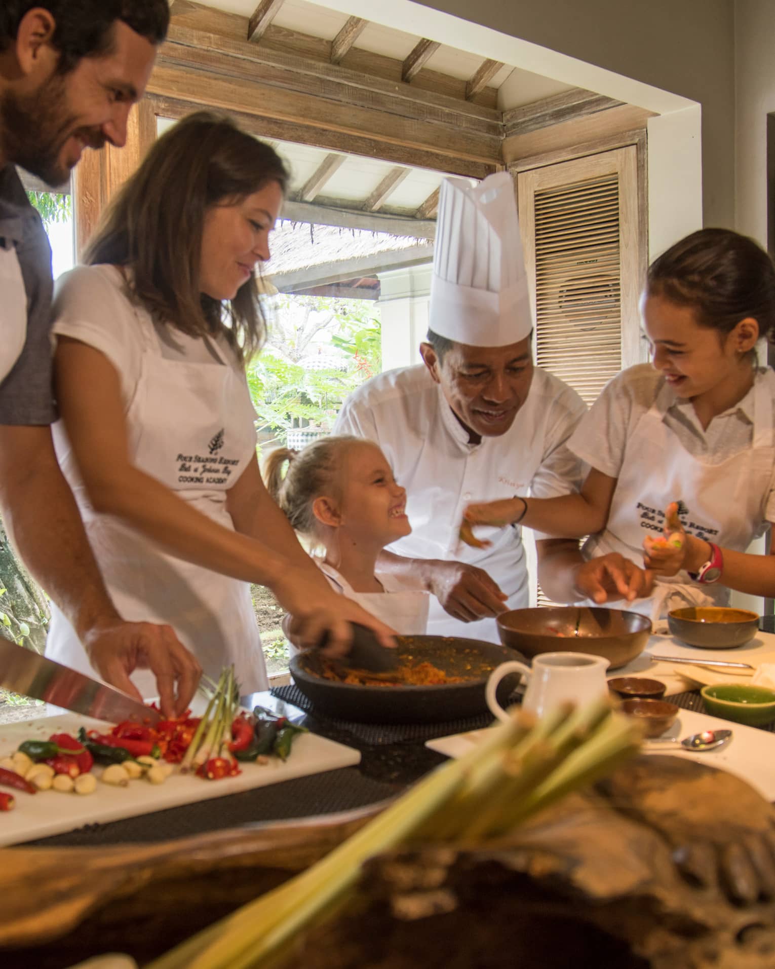 A smiling chef oversees a family joyfully indulging in a private cooking class at Four Seasons