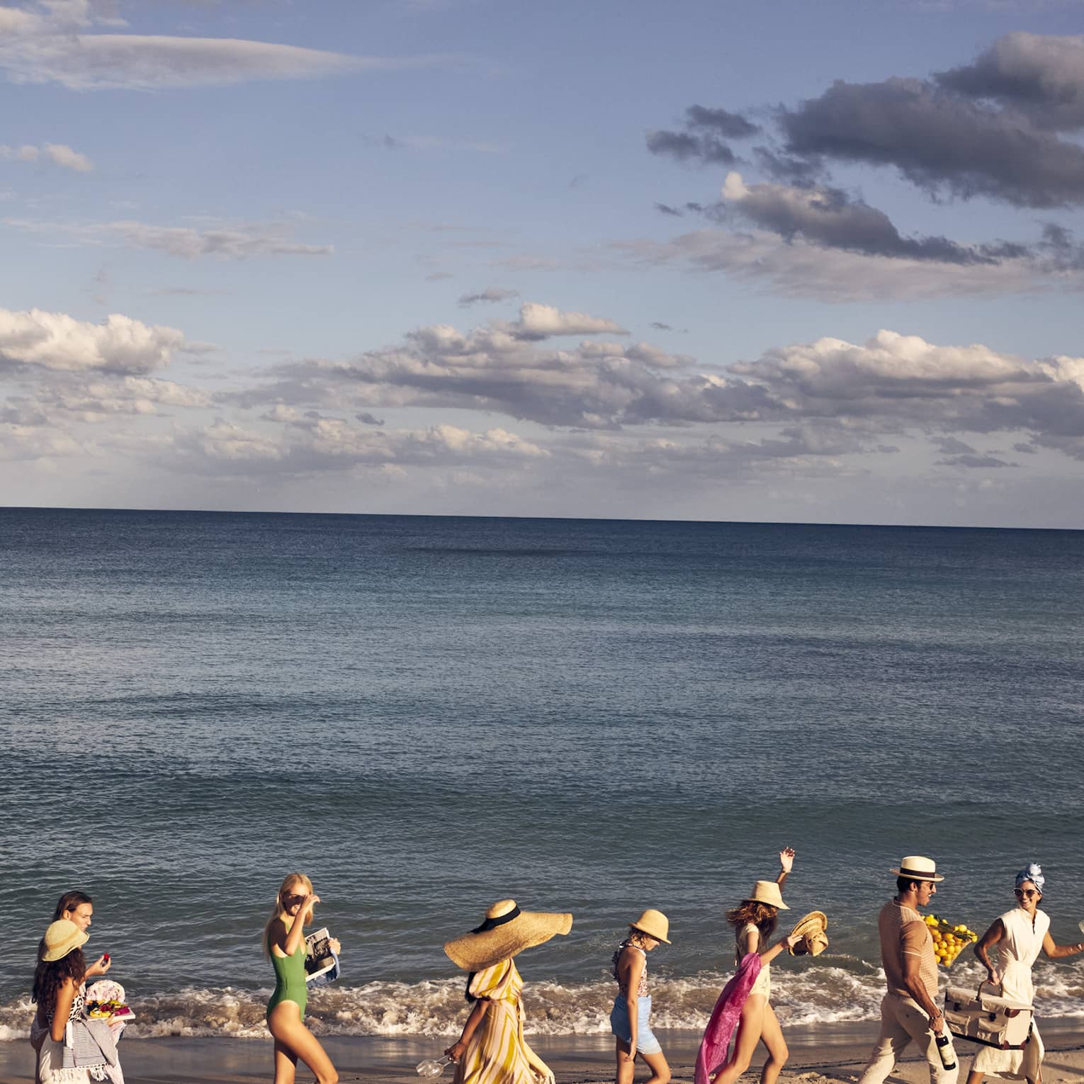 A group of people walking along a sandy beach by the ocean, wearing summer clothing and hats, with a backdrop of a calm sea and partly cloudy sky.