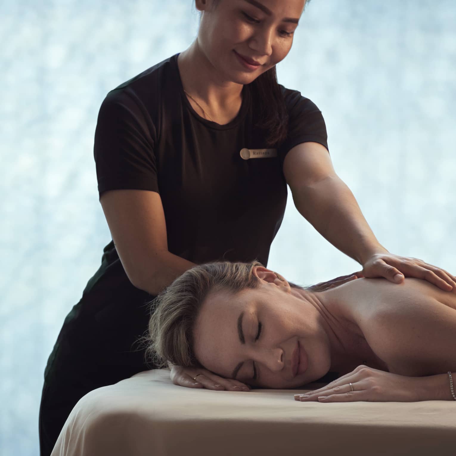 Spa staff massages woman's shoulders as she lays on bed in dark room