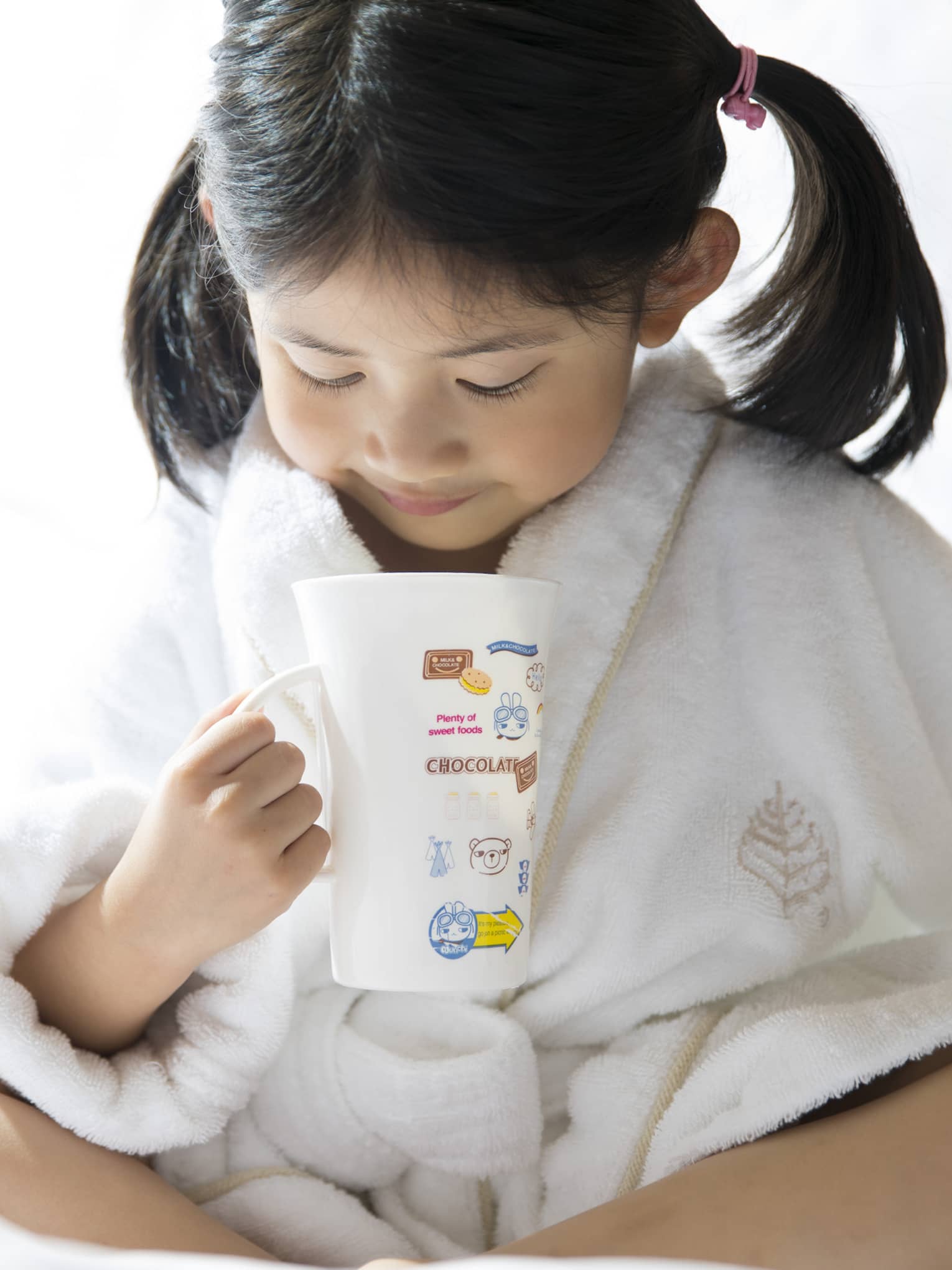 Young child in white bathrobe with hair in ponytails sits cross-legged on bed, holds mug of hot chocolate