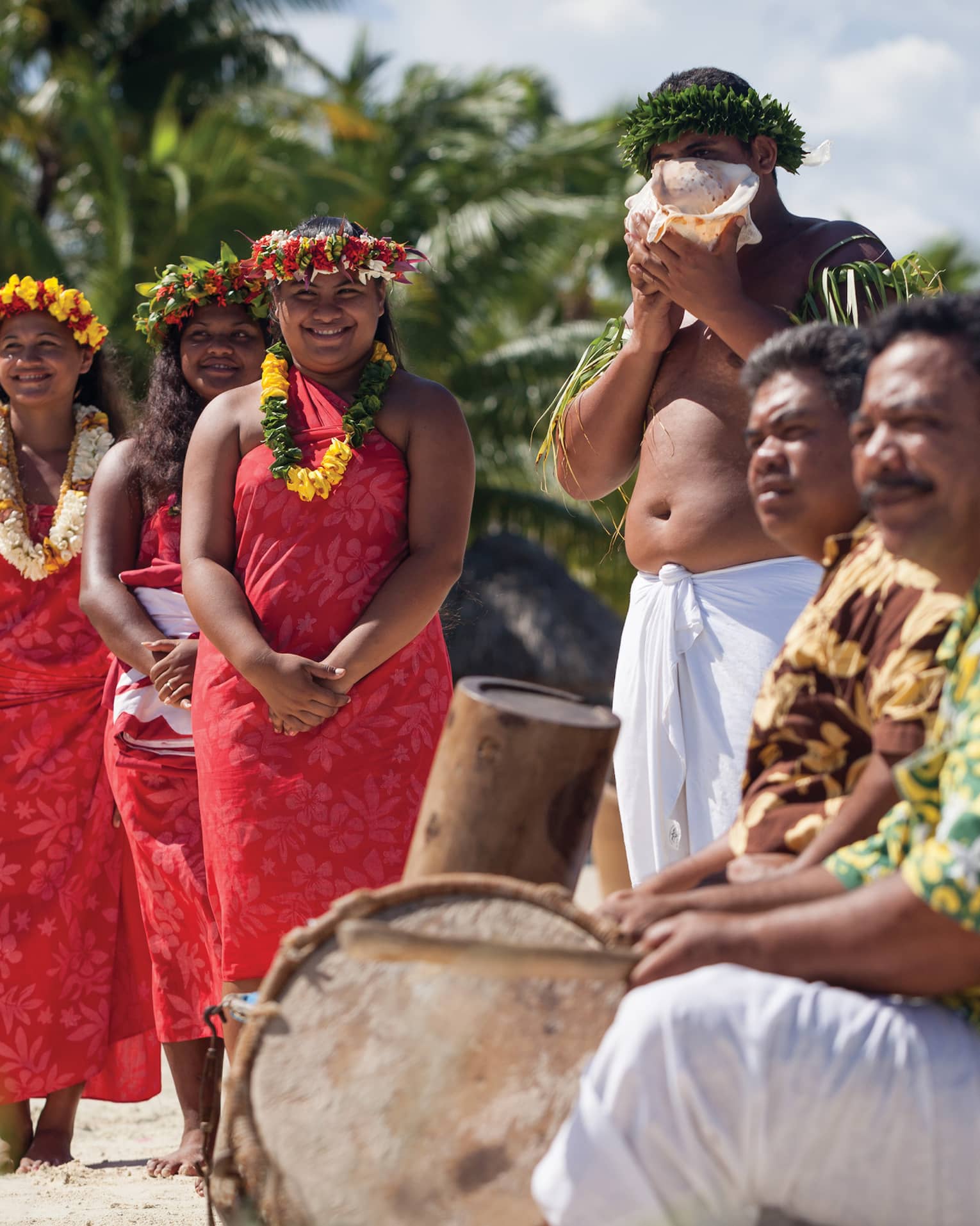 Group of people in traditional attire with leis and a conch shell, standing on a beach