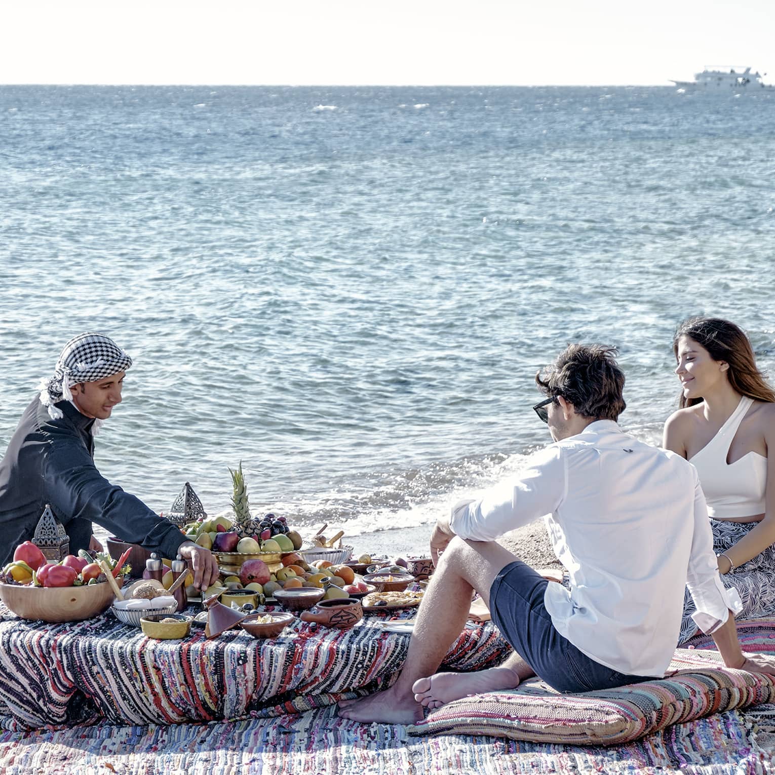 Three people sit on a beach around a low table set with a luxurious array of fruit, bread, and clay dishes full of food.