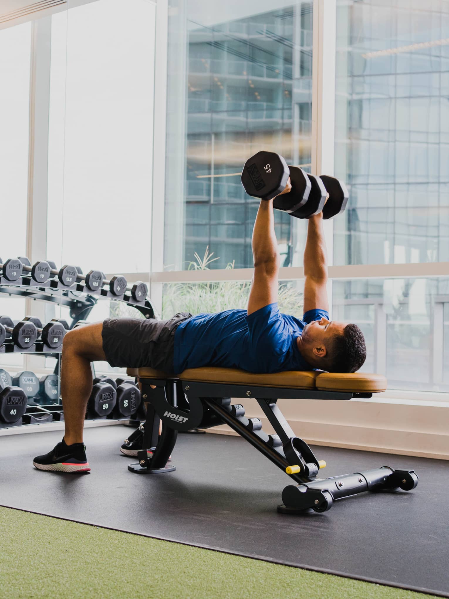A man working out with free weights in a clean fitness room with large windows.