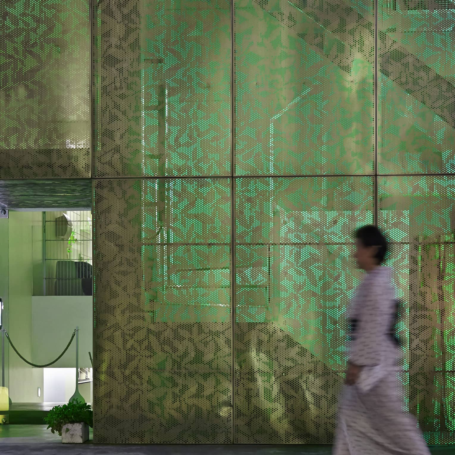 A person heads toward a door to a modern room, passing a multi-paned screened wall with a leaf pattern illuminated in green.