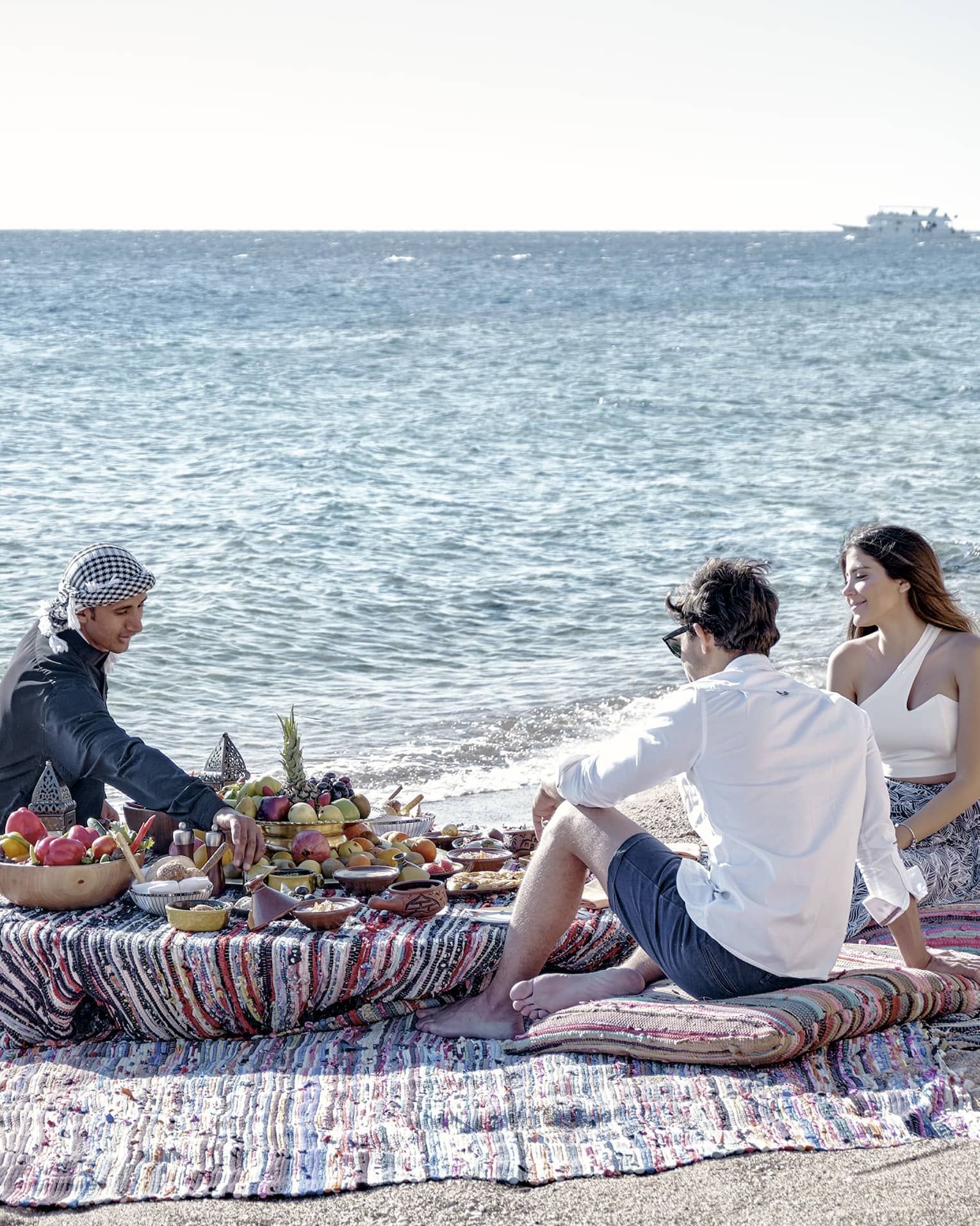 Three people sit on a beach around a low table set with a luxurious array of fruit, bread, and clay dishes full of food.