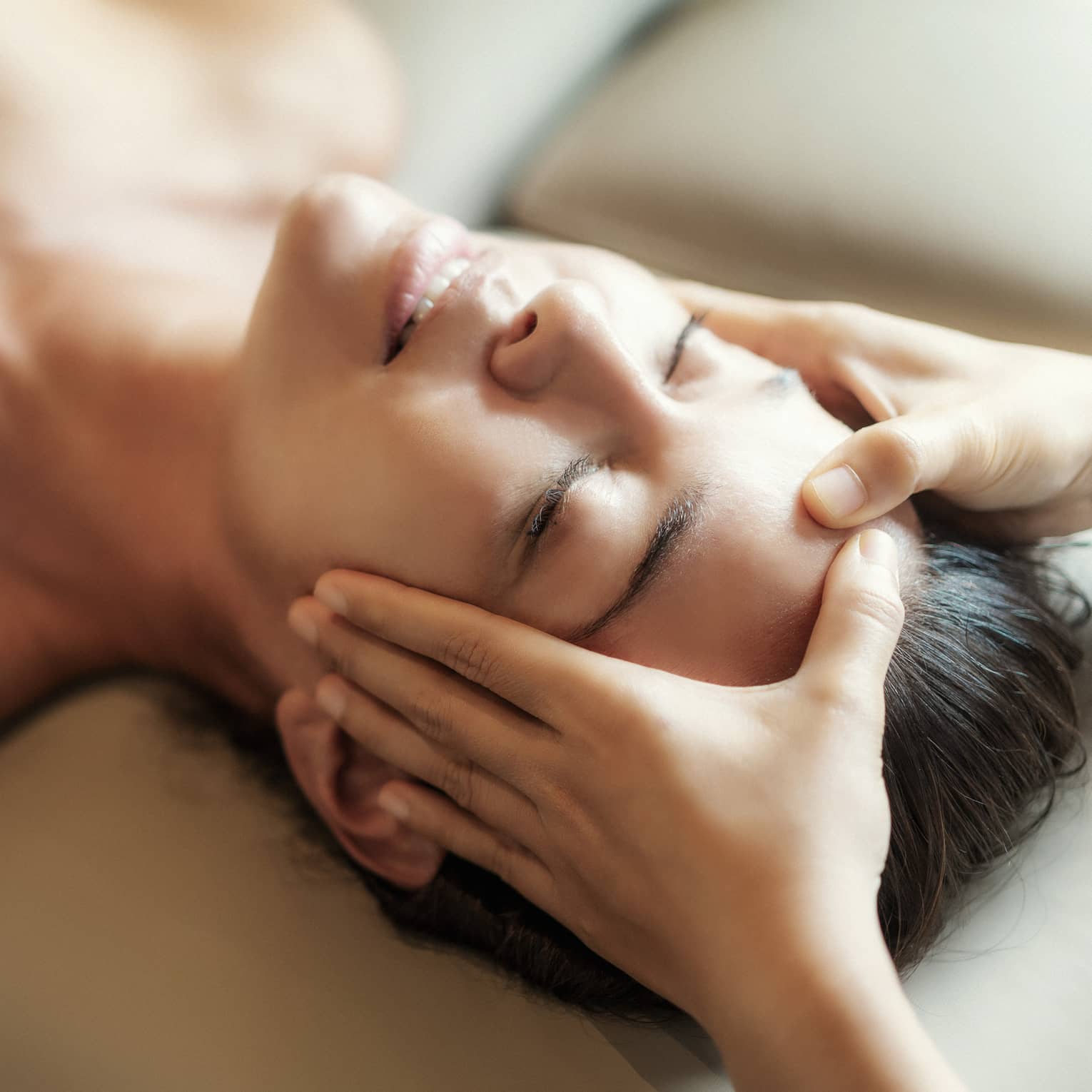 Spa facial, two hands rest on woman's forehead as she closes her eyes, lays under sheet on treatment table