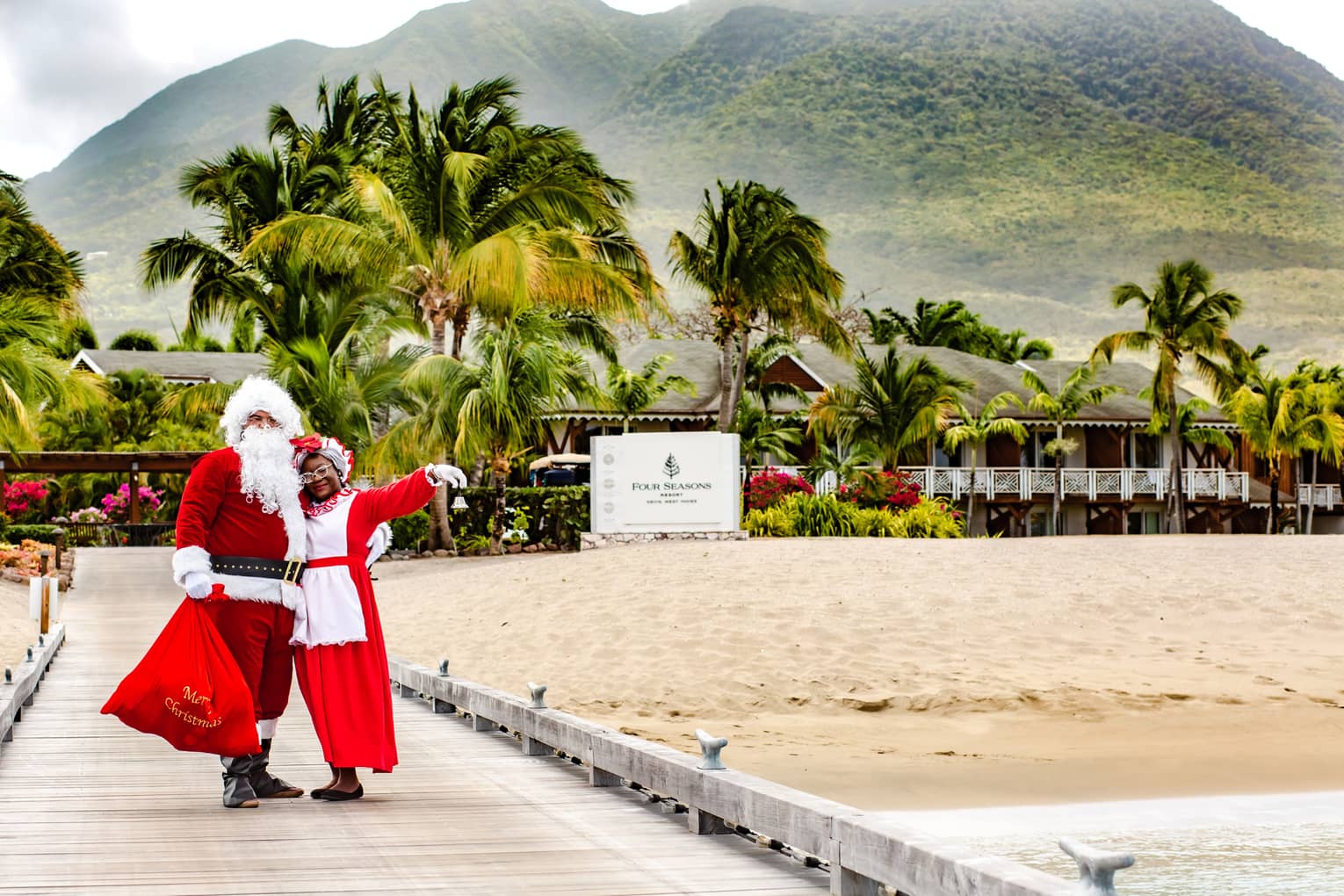 Couple dressed up as Santa and Mrs. Claus stand in front of Resort sign, mountains and palm trees visible in background