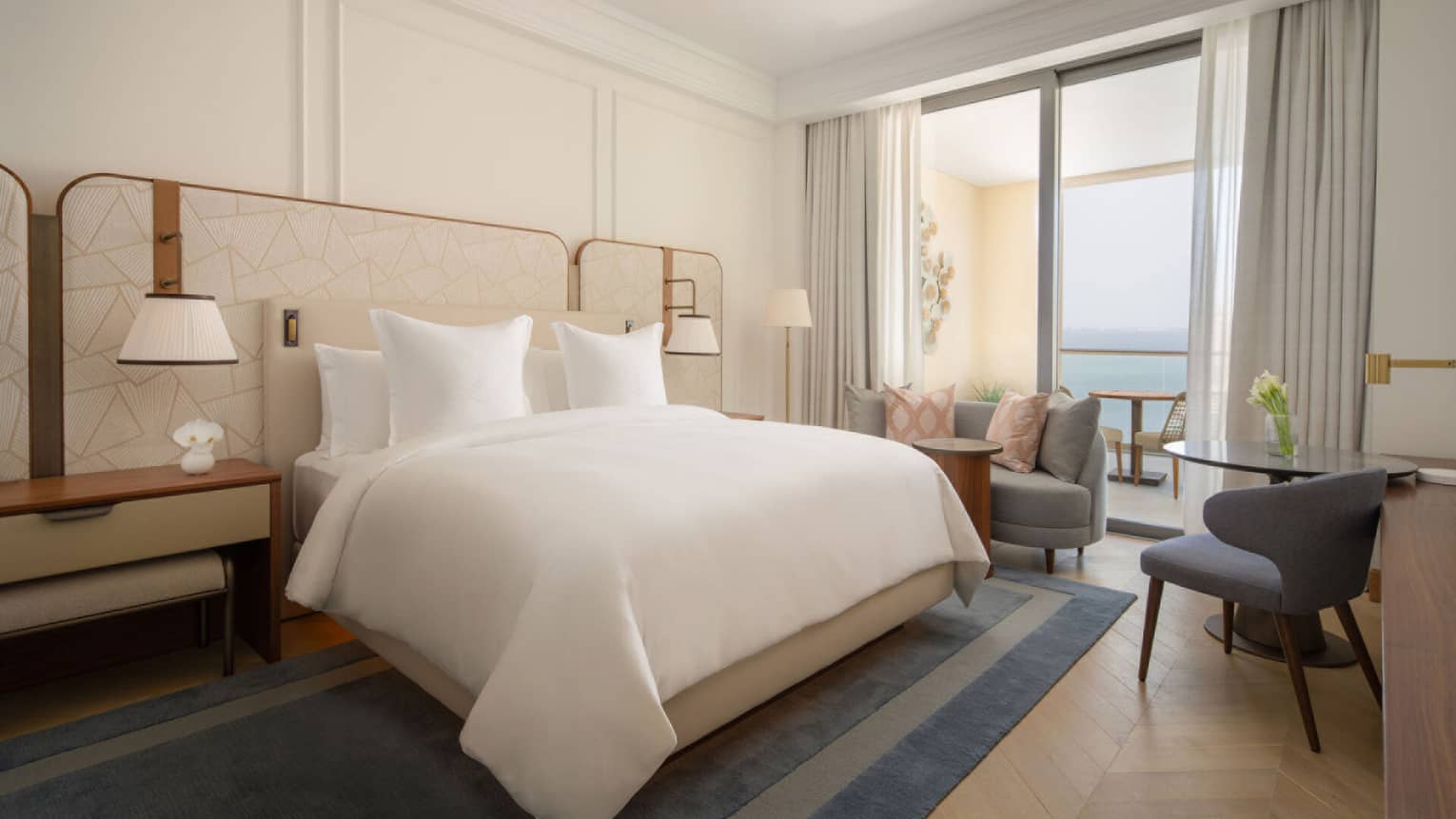 Bright bedroom with a large bed, neutral tones, balcony seating area and sea view through sliding glass doors.