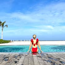 A person in a red swimsuit sits at the edge of a pool, gazing at the ocean under a clear blue sky, creating a serene and picturesque scene.