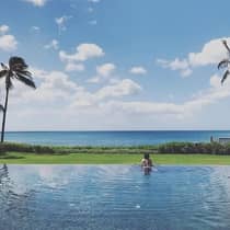 Woman wades in water at edge of swimming pool, looks out at green lawn, ocean