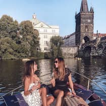 Two people sitting on a boat, enjoying champagne, with a wicker picnic basket and historic buildings and a bridge in the background.