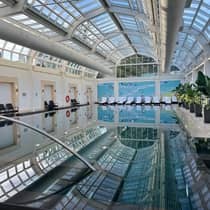 A large indoor pool with a glass ceiling above, surrounded by lounge chairs and green plants