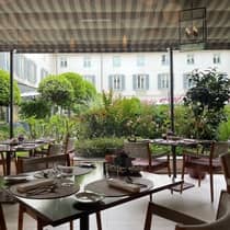 Several dining tables are set with silverware and glasses underneath a fabric covering on an outdoor terrace surrounded by greenery, at the Four Seasons Hotel Milan