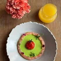 Aerial view of a wooden table with a pink floral arrangement, glass of orange juice and a colourful tomato dish served with green gazpacho