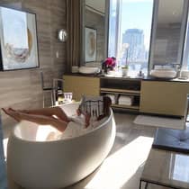 Person reading a magazine while soaking in a freestanding tub in a luxurious bathroom with large windows, double vanity, and city views