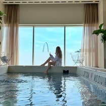 Person in a white robe relaxing by an indoor pool with a glass of champagne, overlooking the Gateway Arch through large windows at a luxury hotel