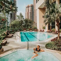 A person in black swimsuit relaxes in a jetted tub at an outdoor pool surrounded by lush greenery and tall residential buildings.