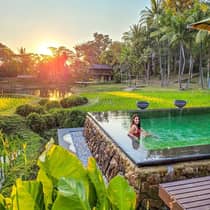 Person relaxing in an infinity pool overlooking rice fields and a pond at sunset, surrounded by lush tropical greenery.