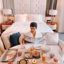 Man wearing bath robe reads newspaper in front of breakfast table, hotel bed