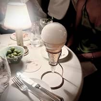 Close-up of a dinner table featuring a unique glass dish with a spherical top, a small lamp, utensils, a bowl of green olives, and drinks in a dimly lit setting.