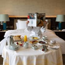 A guest dressed in a white bath robe holds up a Four Seasons magazine behind an in-room dining table topped with a large breakfast spread including waffles, an omelette, pastries, juice and coffee