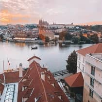 Aerial view of a city with red-roofed buildings along a river, boats on the water and a castle in the background at sunset.