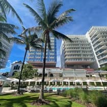 View of a luxurious hotel pool area with tall palm trees, lounge chairs, and umbrellas, framed by modern high-rise buildings under a clear blue sky.