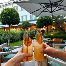 Two hands holding orange cocktails garnished with dried orange slices beneath an umbrella on an outdoor patio at the Four Seasons Hotel Milan