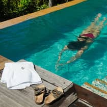 A white towel and pair of sandals sit on the edge of an infinity pool while a woman in a red swimsuit dives underwater