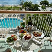 A table filled with colorful breakfast plates, green juice, and coffee on a balcony overlooking the pool and ocean on a sunny day