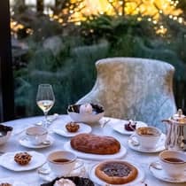 An elegant table set for afternoon tea with a selection of pastries, cakes and cups of tea, surrounded by lush garden and twinkling lights in the background