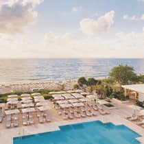 Aerial view of an outdoor swimming pool, rows of chairs, and umbrellas on the deck near the beach and ocean, creating a picturesque seaside escape.