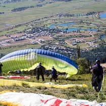Paragliders preparing for takeoff on a grassy slope overlooking a valley with ponds, houses and open fields below